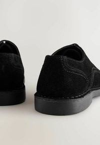 Zapatos de ante negro con punta redondeada, superficie texturizada y una suela de goma negra gruesa. Presenta costuras contrastantes y un interior suave.