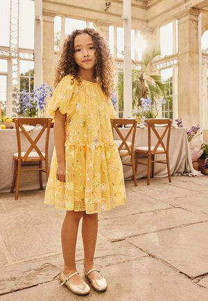 Ung pige med krøllet hår iført en gul blomsterkjole og gyldne sko står i et solbeskinnet rum med træstole og blomster.