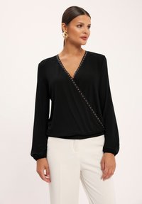 Czarna bluzka z długim rękawem typu wrap z dekoltem w kształcie V i ozdobnymi złotymi ćwiekami wzdłuż dekoltu. Materiał jest gładki z delikatnym spadkiem. Połączona z białymi spodniami.