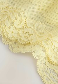 Tissu délicat en dentelle jaune pâle avec motifs floraux et bords festonnés sur un fond clair.