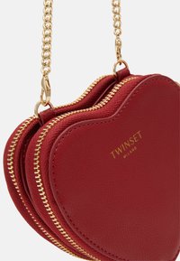 Borsa a forma di cuore in pelle rossa con cerniere dorate e tracolla a catena, con logo del marchio impresso. Texture liscia e design compatto.