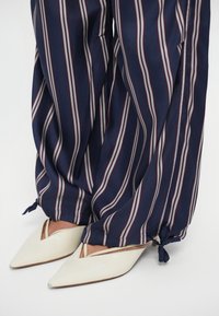 Fehér lakkbőr, hegyes orrú cipők oldalsó kivágásokkal, sötétkék nadrággal párosítva, amely függőleges bordó és szürke csíkokkal, valamint masni díszítéssel rendelkezik az ujjak végén.