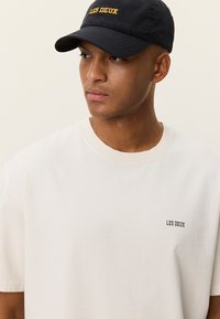 Νεαρός άντρας που φοράει μαύρο καπέλο με την επιγραφή «LES DEUX» και λευκό μπλουζάκι με μικρό λογότυπο «LES DEUX», κοιτάζει προς τα αριστερά σε απλό φόντο.