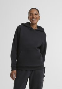 Uśmiechnięta kobieta z kolczykami, ubrana w czarną bluzę z kapturem i ciemne spodnie, stoi na tle jednolitego jasnoszarego tła.