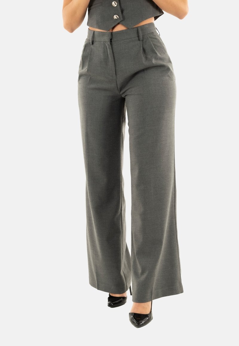 Femme portant un pantalon gris taille haute à jambes larges et des talons noirs pointus, cadrée à la taille avec les mains près du torse.