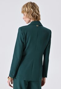 Türaublauer Blazer mit strukturiertem Design, Reverskragen, Ein-Knopf-Verschluss und einem goldenen Akzent auf der Rückseite in der Nähe des Kragens. Lange Ärmel.