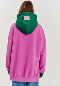 Prekinsicová mikina vo veľkosti oversize v ružovej flísovej látke so zelenou kapucňou. Na zadnej strane sa nachádza ružová nášivka. Rukávy a lem sú rebrované.
