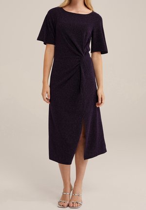 Nainen, joka pukeutunut tummanpurppuraiseen, teksturoituun midimekkoon, jossa on lyhyet hihat, sivusolmuke ja edessä halkio, yhdistettynä hopeisiin remmikenkiin.