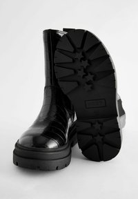 Schwarze Knöchelstiefel aus glänzendem Krokodil-Präge-Material. Mit einer klobigen schwarzen Gummisohle mit Sternmuster-Profil und Zuglasche.