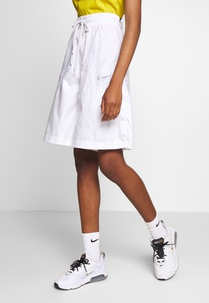 Personne portant un short blanc à hauteur des genoux, des baskets Nike blanches, des chaussettes blanches et un haut jaune sur un fond clair uni.