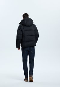 Schwarze Daunenjacke mit Kapuze, mit einem gesteppten Design, getragen über blauen Jeans und braunen Stiefeln. Minimalistischer Stil und kompakte Form.