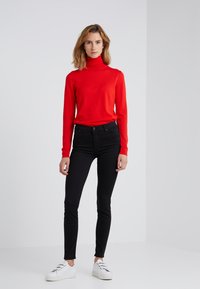 Pull à col roulé rouge, style ajusté, associé à un jean skinny noir et des baskets blanches. Texture lisse, design minimaliste, sans motifs visibles.