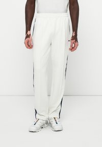 Pantalones de chándal blancos con cinturilla elástica y franjas laterales en azul marino. Tela suave con un corte relajado, combinados con zapatillas blancas.