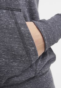 Heatherad mörkgrå hoodie-tyg med sidficka, med texturerad stickning och förstärkta sömmar längs kanterna.