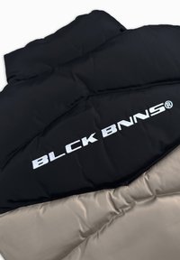 Puffermantel mit schwarzem Oberteil und beigem Unterteil, mit auffälligem reflektierendem "BLCK BNNS"-Logo in Weiß auf der Vorderseite.