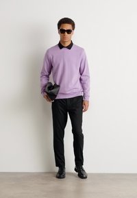 Ανοιχτό μωβ πουλόβερ με μαύρο γιακά, μαύρο παντελόνι και μαύρα παπούτσια. Μοντέλο που κρατάει μια μαύρη τσάντα χειρός, φοράει γυαλιά ηλίου. Απλό φόντο.