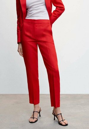 Nainen pukeutuneena punaisiin räätälöityihin housuihin ja vastaavaan bleiseriin, valkoinen paita, seisoo harmaalla lattialla, mustat remmikkäät korkokengät näkyvissä.