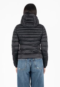 Zwarte pufferjack met een capuchon, voorzien van gequilte delen en elastische manchetten. Draagt over een blauwe spijkerbroek, wat een aansluitende silhouette laat zien.