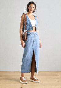 Veste en denim cropped, chemise blanche et longue jupe en denim bleu avec une fente à l'avant. Sac crossbody marron et baskets marron à motifs.
