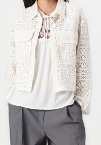 Biała koronkowa krótka kurtka z misternymi kwiatowymi wzorami, dwoma kieszeniami na piersi i okrągłym kołnierzykiem. Nosi się ją na białej bluzce i szarych spodniach.