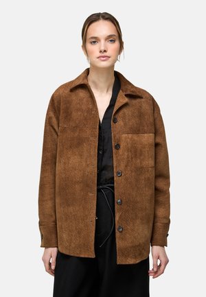 Jonge vrouw die een bruine, getextureerde jasje met knopen draagt over een zwart shirt en zwarte broek, staand tegen een witte achtergrond.