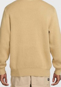 Beige långärmad stickad tröja bärs av en person med synliga händer och beiga byxor, sedd bakifrån mot en enkel ljus bakgrund.