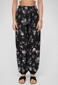 Femme portant un pantalon fluide noir à motifs floraux avec une taille élastique et des poignets, associé à des sandales noires à lanières croisées et un crop top blanc.