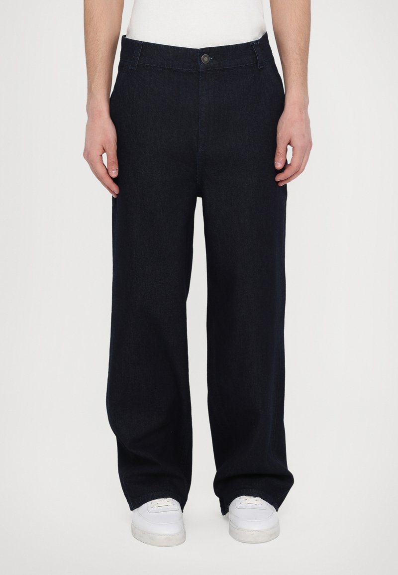 Dark blue denim trousers with a straight leg design, featuring a front button closure and side pockets, paired with white sneakers.