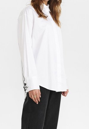 Femme portant une chemise blanche ample à boutons avec un dessin visage en ligne noire sur le poignet de la manche et un pantalon noir.