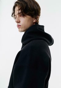 Giovane uomo con capelli castani di lunghezza media che indossa una felpa nera con cappuccio, che guarda a sinistra su uno sfondo bianco semplice.