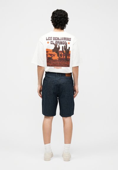 Άτομο με κατσαρά μαλλιά φοράει λευκό T-shirt με πορτοκαλί γραφικό σχεδιασμό ενός καουμπόη πάνω σε άλογο και κάκτο, με το κείμενο «Les Benjamins El Gringo», και σκούρα σορτς με λεπτές ρίγες.