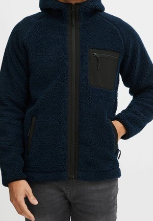 Homme portant une veste en polaire sherpa bleu marine avec fermeture éclair noire et poches sur la poitrine et les côtés avec fermeture éclair, une main dans la poche.