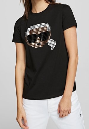 Osoba ubrana w czarną koszulkę z pikselową grafiką twarzy w okularach przeciwsłonecznych oraz białe spodnie, stojąca na neutralnym tle.