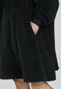 Coperta con cappuccio nera, di grande dimensione, realizzata in tessuto pile morbido. Presenta ampie tasche frontali e polsini a costine. Lunghezza fino a metà coscia.