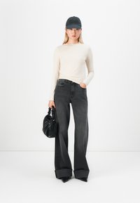 Ungdommelig kvinne iført en beige langermet topp, svart vide bukser med oppbrett, svart caps, holder en svart håndveske og står mot en hvit bakgrunn.