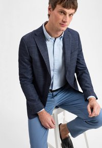 Marineblau strukturierter Blazer mit zwei Knöpfen, getragen über einem hellblauen Hemd, kombiniert mit hellblauen Hosen und schwarzen Slippern.