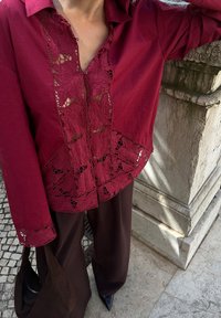 Blusa bordeaux con inserti in pizzo floreale e chiusura con bottoni, abbinata a pantaloni larghi marroni e una borsa scura, su uno sfondo di pietra.
