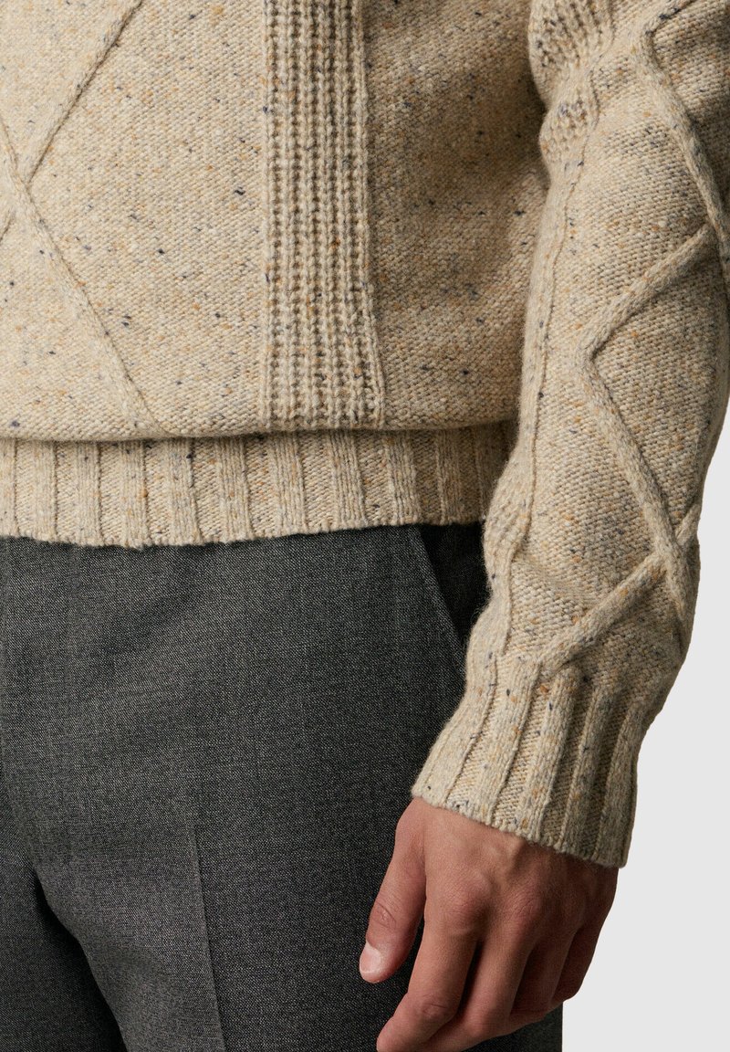 Beżowy sweter dzianinowy z wzorami warkoczowymi i prążkowanymi na rękawie i dole, zestawiony z ciemnoszarymi teksturowanymi spodniami, pokazujący swobodną pozycję dłoni.