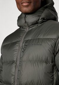 Casaco puffer verde escuro com gola alta, design acolchoado horizontal, fecho éclair e logótipo bordado no peito. Textura suave.