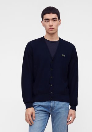 Junger Mann trägt eine dunkelblaue, geknöpfte Strickjacke über einem schwarzen Hemd und hellblauen Jeans und posiert vor einem einfarbigen weißen Hintergrund.
