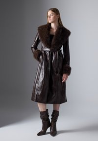 Cappotto in pelle marrone con colletto e polsini in pelliccia sintetica, vita con cintura, lunghezza al ginocchio, caratterizzato da una texture a coccodrillo. Modella che indossa stivali alti coordinati.