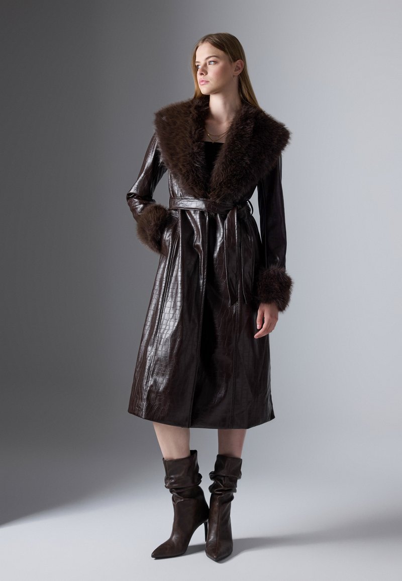 Cappotto in pelle marrone con colletto e polsini in pelliccia sintetica, vita con cintura, lunghezza al ginocchio, caratterizzato da una texture a coccodrillo. Modella che indossa stivali alti coordinati.