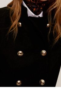 Sort dobbeltradet frakke med store guldknapper og høj krave, parret med et mønstret sort og orange tørklæde.