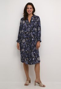 Rochie bleumarin cu un model floral și paisley, mâneci lungi, decolteu în V și o croială lejeră. Asortată cu sandale deschise la vârf, bej.