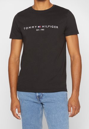 T-shirt en coton noir avec col rond, arborant le texte logo blanc "TOMMY HILFIGER EST. 1985" centré sur la poitrine. Manches courtes, coupe décontractée.