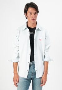 Lys blå denimskjorte med knapper, lange ermer oppbrettet, brukt over en svart skjorte. Merkbar rød logo på venstre bryst.