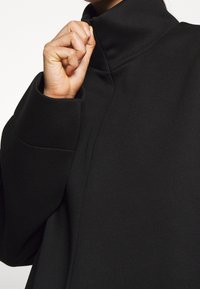 Černý kabát s vysokým límcem vyrobený z hladké, strukturované látky. Má zapínání na zip na límci a široké rukávy pro uvolněný střih.