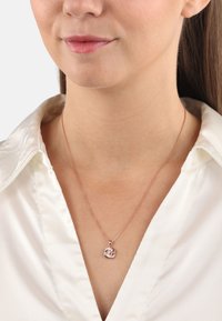 Collana con pendente in oro rosa su una catena sottile, caratterizzata da un design a vortice e un accento scintillante, indossata sopra una blusa in satin bianco.