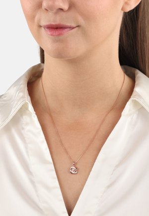 Collana con pendente in oro rosa su una catena sottile, caratterizzata da un design a vortice e un accento scintillante, indossata sopra una blusa in satin bianco.
