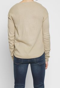 Beżowy prążkowany sweter z okrągłym dekoltem i opuszczonymi szwami na ramionach, zestawiony z ciemnymi dżinsami z ozdobnym przeszyciem na kieszeniach z tyłu.